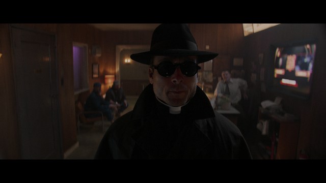 A scene from a film featuring two characters, one elderly woman holding a small object and speaking animatedly, and a young man listening intently, surrounded by lush greenery and plants in a greenhouse setting.