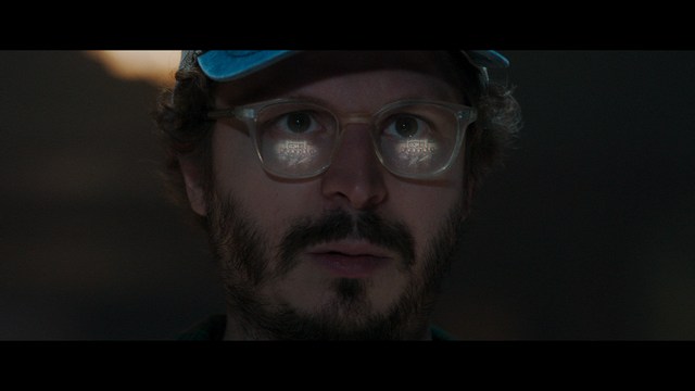 A scene from a film featuring two characters, one elderly woman holding a small object and speaking animatedly, and a young man listening intently, surrounded by lush greenery and plants in a greenhouse setting.