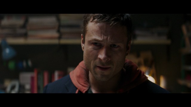 A scene from a film featuring two characters, one elderly woman holding a small object and speaking animatedly, and a young man listening intently, surrounded by lush greenery and plants in a greenhouse setting.