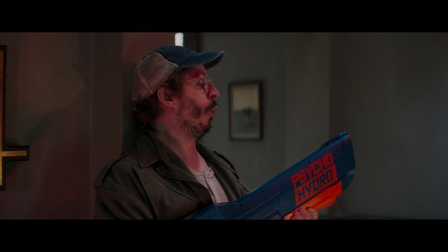 A scene from a film featuring two characters, one elderly woman holding a small object and speaking animatedly, and a young man listening intently, surrounded by lush greenery and plants in a greenhouse setting.