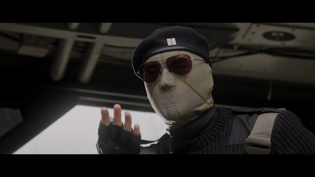 A scene from a film featuring two characters, one elderly woman holding a small object and speaking animatedly, and a young man listening intently, surrounded by lush greenery and plants in a greenhouse setting.