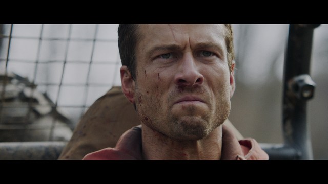 A scene from a film featuring two characters, one elderly woman holding a small object and speaking animatedly, and a young man listening intently, surrounded by lush greenery and plants in a greenhouse setting.