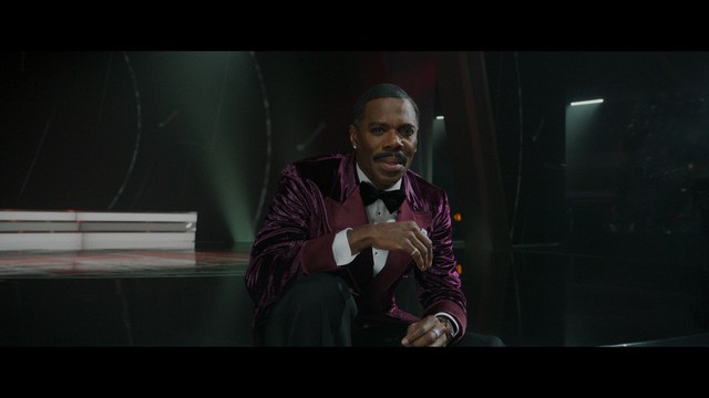 A scene from a film featuring two characters, one elderly woman holding a small object and speaking animatedly, and a young man listening intently, surrounded by lush greenery and plants in a greenhouse setting.