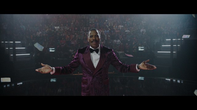 A scene from a film featuring two characters, one elderly woman holding a small object and speaking animatedly, and a young man listening intently, surrounded by lush greenery and plants in a greenhouse setting.