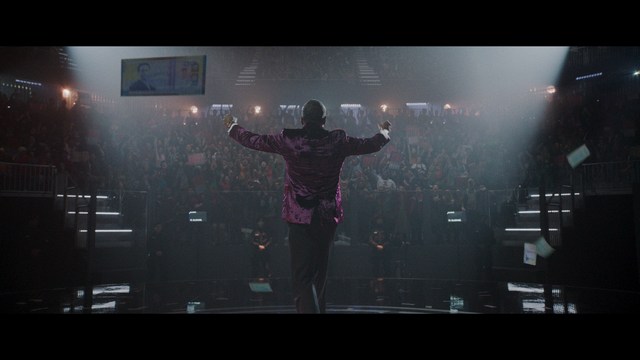 A scene from a film featuring two characters, one elderly woman holding a small object and speaking animatedly, and a young man listening intently, surrounded by lush greenery and plants in a greenhouse setting.