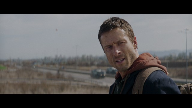 A scene from a film featuring two characters, one elderly woman holding a small object and speaking animatedly, and a young man listening intently, surrounded by lush greenery and plants in a greenhouse setting.