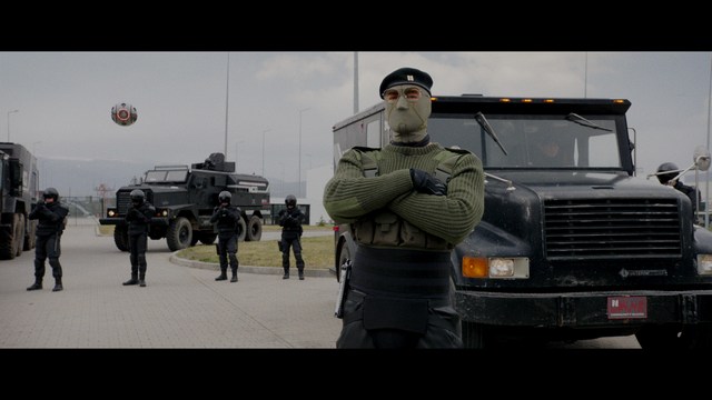 A scene from a film featuring two characters, one elderly woman holding a small object and speaking animatedly, and a young man listening intently, surrounded by lush greenery and plants in a greenhouse setting.