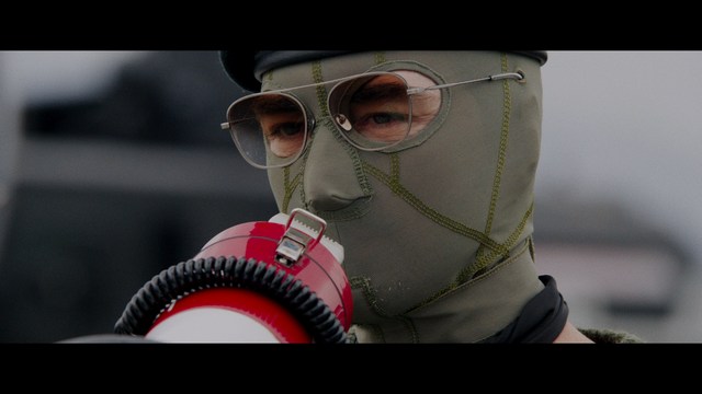 A scene from a film featuring two characters, one elderly woman holding a small object and speaking animatedly, and a young man listening intently, surrounded by lush greenery and plants in a greenhouse setting.