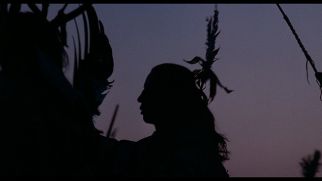 A still from a movie featuring a man standing on a boardwalk in front of an arcade with signs for Skeeball, saltwater taffy, and various attractions.