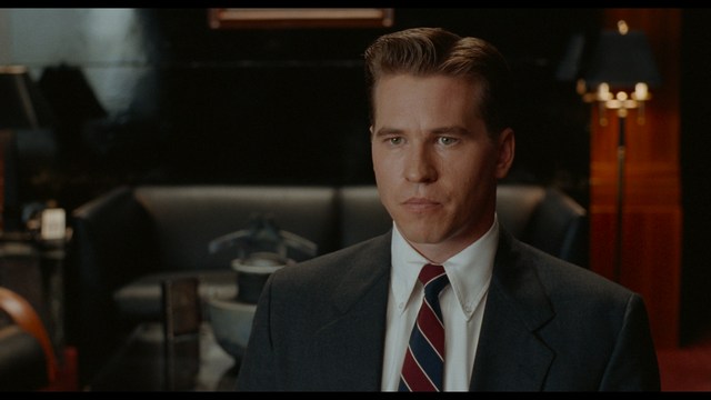 Statue of a smiling figure resembling Jesus, giving a thumbs up gesture, set against a colorful backdrop with signs related to Catholicism.