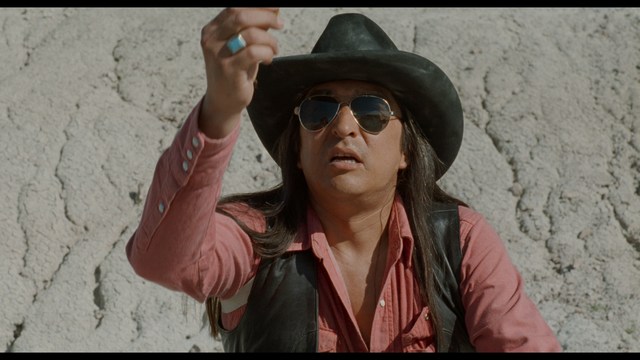 A man in a white suit and hat stands at a doorway, smiling, with a suburban landscape in the background.