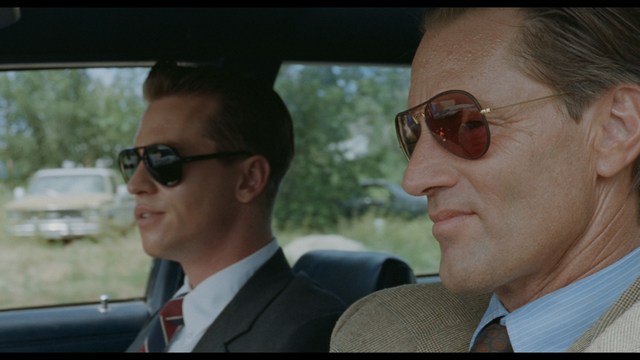 A man with large, feathered angel wings stands in a room, wearing a black suit and looking directly at the viewer.