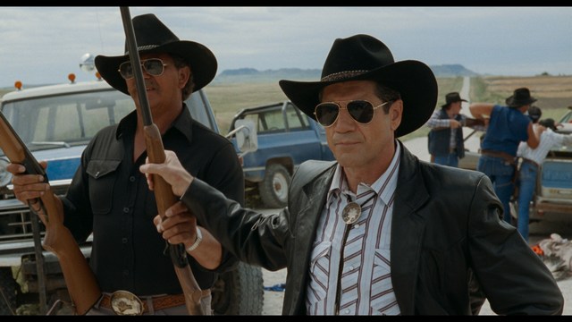 A close-up of a man with a concerned expression, with blurred figures in the background, set in a dimly lit interior.