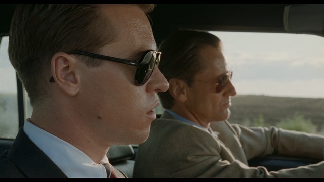 Two characters sitting in a diner, one wearing a blue beanie with 'SNOOGANS' and the other wearing a cap, both looking intently at something off-screen.