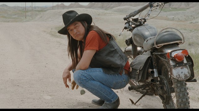 A scene featuring a man with short hair and a beard, wearing a brown jacket and purple shirt, standing outside a restaurant with bright yellow walls and a cartoon cow logo.