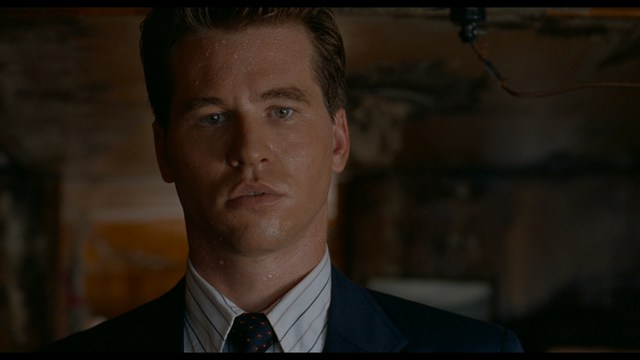 Close-up of a woman with long black hair sitting in a restaurant, looking concerned. A colorful painting is visible in the background, along with red bar stools.