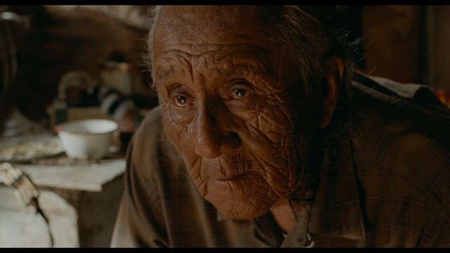 Two young men are looking at something off-camera, standing inside a building with bright lighting and seating areas visible in the background.