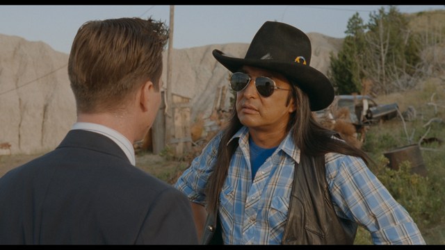 A close-up shot of a man wearing a white suit and a white hat, looking intently at someone in a retail store filled with merchandise.