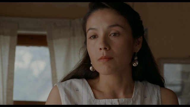 Close-up of a man with a serious expression, seated in a dimly lit area, with soft lighting from background lamps.