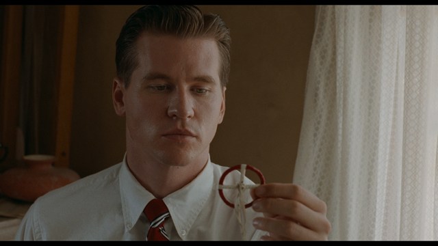 A close-up shot of a young man with short hair and a gray sweatshirt, looking intently at the camera, with a woman sleeping beside him in the background.