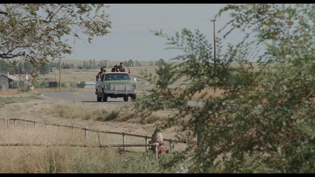 A woman seated at a restaurant table, thoughtfully resting her chin on her hand while reading a newspaper. Blurred figures of two people can be seen in the background.