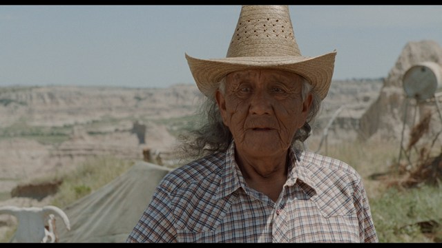 A man with dark, styled hair and a subtle smile seated at a restaurant table, with blurred diners in the background.