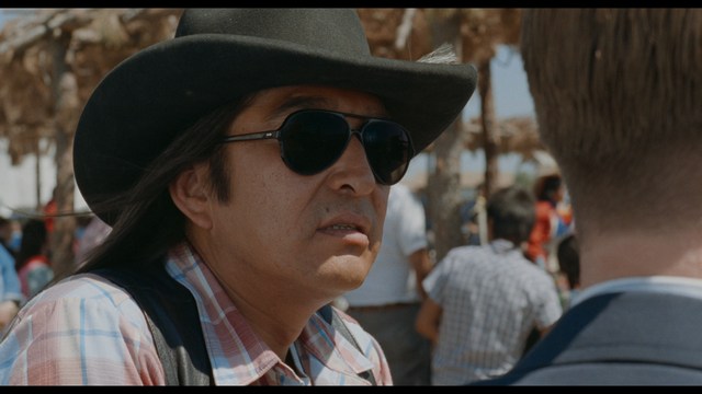 Two characters sitting at a table, one holding a small statue, with a surprised expression. The background features vibrant colors and patterns.