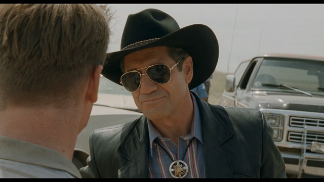 Two figures walking on a road towards a blue 'Welcome to New Jersey' sign.