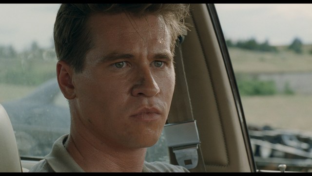 A scene featuring a man in a bishop's attire speaking at a podium surrounded by microphones, with a woman in a red dress beside him and a crowd of onlookers in the background.