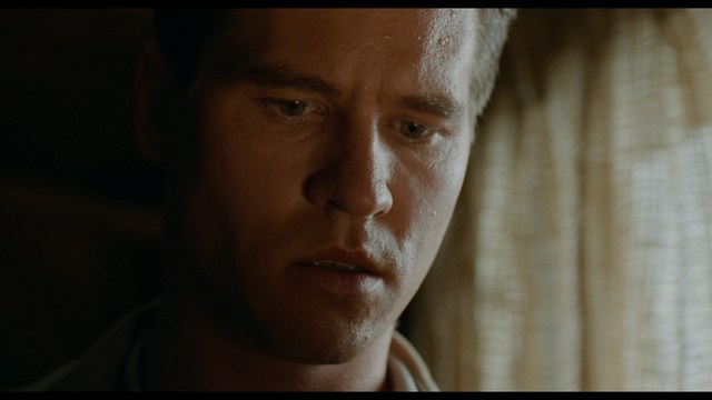 A tense scene featuring a woman with long hair and a serious expression, looking off to the side, while a man with short hair sits in the background, appearing relaxed and slightly turned away.