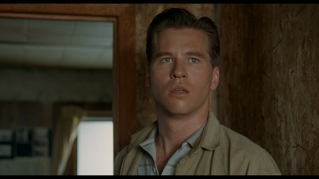 A shocked man with long hair and a baseball cap sitting in a bar, looking surprised.