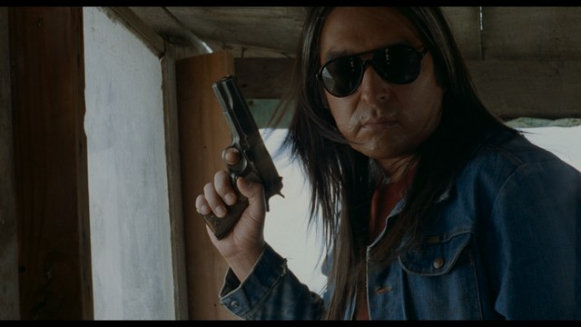 A man with long hair and sunglasses is holding a gun while standing in a dimly lit area, looking intently outwards.