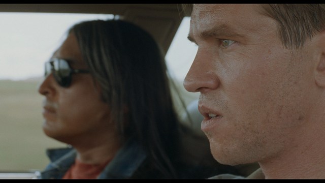 A close-up shot of two men sitting in a car, one wearing sunglasses and the other with a concerned expression, in a scenic landscape.