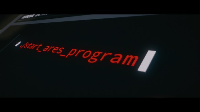 A man with a beard and mustache smiles warmly while standing on a boardwalk by the beach, with colorful flags overhead and a large building in the background.