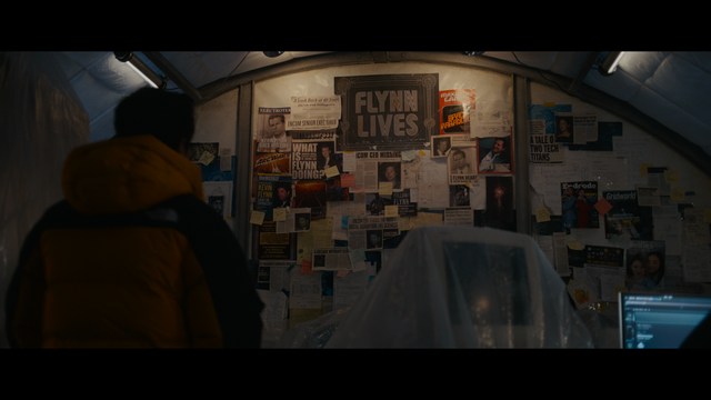 A woman with a pensive expression rests her chin on her hand, sitting at a table with kitchen appliances and items in the background.
