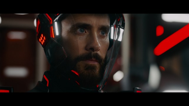 A man with large, feathered angel wings stands in a room, wearing a black suit and looking directly at the viewer.