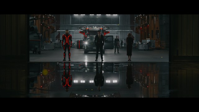 A close-up of a man with a concerned expression, with blurred figures in the background, set in a dimly lit interior.