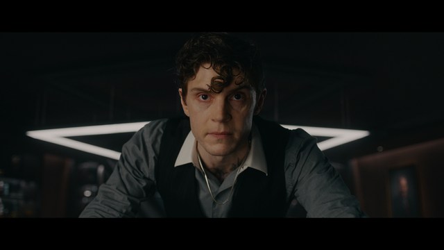 Close-up of a man with a serious expression, seated in a dimly lit area, with soft lighting from background lamps.