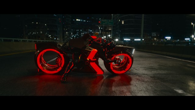 A close-up shot of a young man with short hair and a gray sweatshirt, looking intently at the camera, with a woman sleeping beside him in the background.