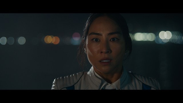 A serious-looking cardinal with a red hat and black robes sitting in a wooden chair in an office, surrounded by colorful posters and memorabilia.