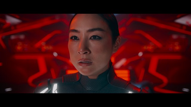 A scene featuring a man in a bishop's attire speaking at a podium surrounded by microphones, with a woman in a red dress beside him and a crowd of onlookers in the background.