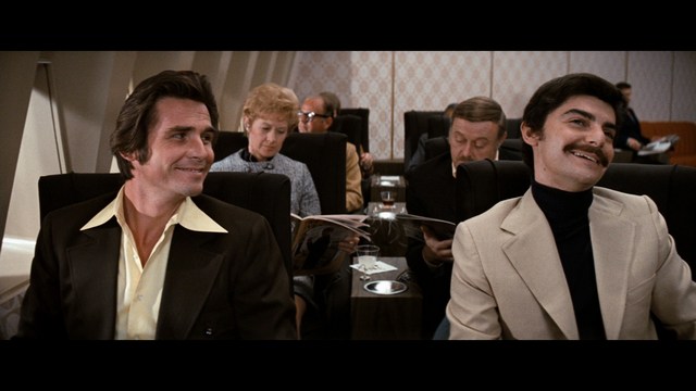 A smiling man with a beard stands on a wooden boardwalk near the beach, with colorful flags in the background and a building visible in the distance.