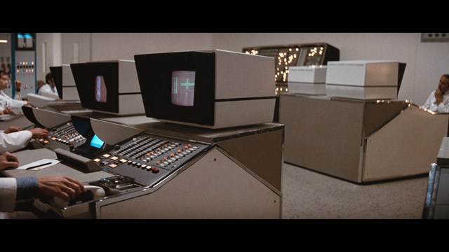 Two male characters sitting in a waiting area, one wearing a black jacket and the other in a maroon hoodie, with popcorn in hand.