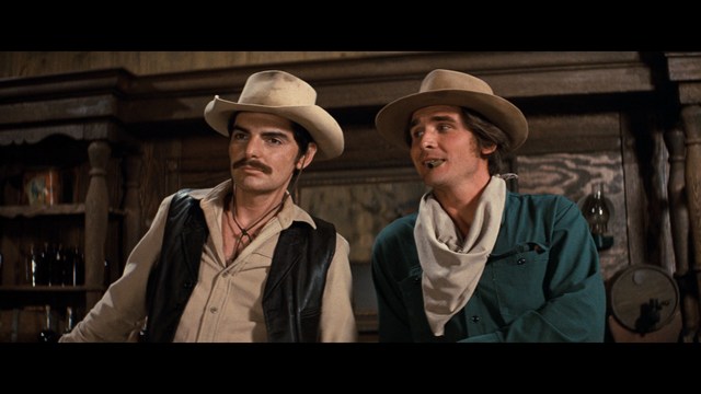 A woman with long dark hair, wearing a brown sweater, gestures with one hand while speaking in a warmly lit room with blinds and colorful artwork in the background.