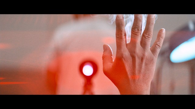 A man with large white wings stands in a room, wearing a dark suit, with a serious expression on his face. Bookshelves and curtains are visible in the background.