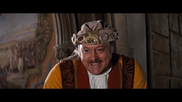 A man with short hair wearing a brown jacket and purple shirt, speaking animatedly in front of a colorful restaurant featuring a cartoon cow logo in the background.