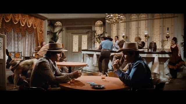 A scene from a movie featuring a man with short black hair and a dark jacket, sitting at a table with a surprised expression, holding a beverage, in a dimly lit indoor setting.