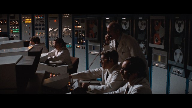 Close-up shot of a young man with short hair, wearing a gray hoodie, looking over his shoulder with a contemplative expression, while another person is asleep in the background.