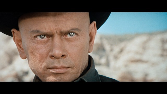 Two male characters in a cozy indoor setting, one with long hair and a beanie, smirking while holding a small object, and the other with a beard and cap, looking surprised. A colorful background with posters can be seen.