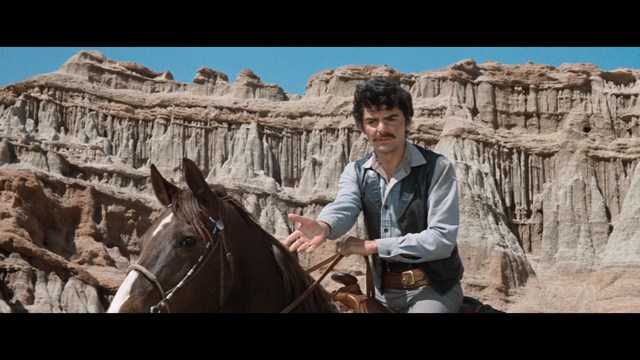 A close-up of a man wearing a red cardinal's hat and black clerical robes, seated in a leather armchair with a serious expression, in an office setting adorned with colorful posters.